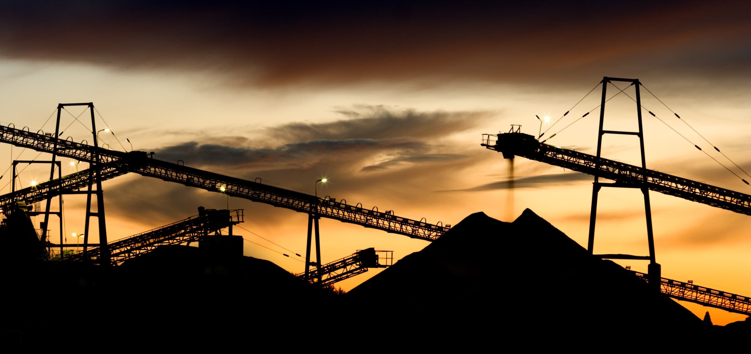 Silhouetted conveyor gantries and a stockpile against an orange sunset sky